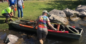 People pulling a boat ashore that is filled with marine debris collected along a lake shoreline.