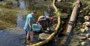 Two people are standing in a shallow creek on a sunny day cleaning out litter from a yellow litter boom that is stretched across the channel.
