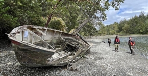 A broken wooden boat lays on it side on a rocky, tree-lined shoreline with three government officials in the background. 