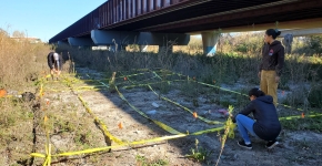 Three researchers use yellow tape to set up a grid covering bare and grassy vegetation for observing marine debris via uncrewed aerial systems near a railroad bridge overpass.