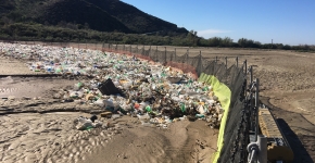 A large trash boom goes from one side of a dry river bed to the other with lots of trash stuck on one side.