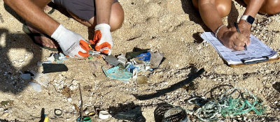 Two people's hands are seen examining and documenting small plastic pieces and netting on a data card on a sandy beach.