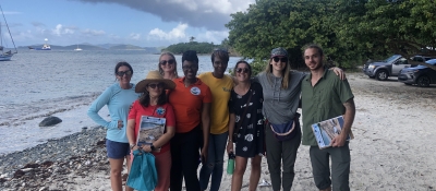 A group of eight people stand on a U.S. Virgin Island beach with clipboards. 