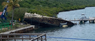 A derelict vessel grounded on its side on a shoreline near a dock.