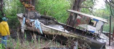 One person stands and looks at a boat that is being pulled from the water with an excavator.