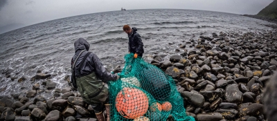 Two people drag a mass of derelict fishing nets, buoys, and other gear on a rocky Alaska shoreline.