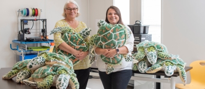 Two people hold life size stuffed turtles.