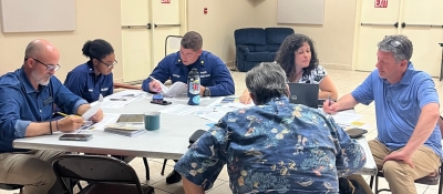 A group of six people sit around a table talking and working with laptops and paper materials.