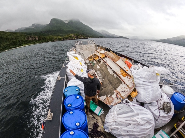 A person pointing towards a mountainous shoreline aboard a moving boat that is full of various marine debris items and a derelict vessel.