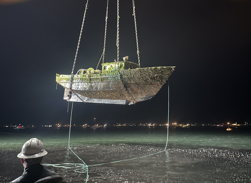 A derelict vessel being hoisted out of the water with a series of chains at night with a city skyline illuminated in the distance.