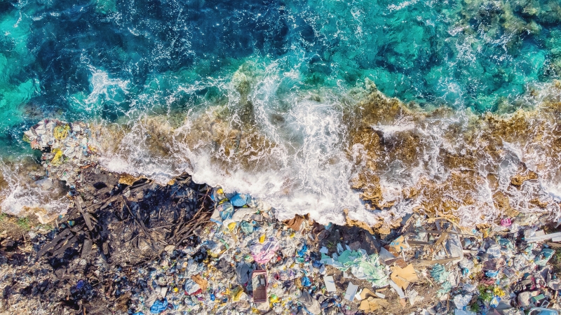 An aerial photograph of a wave crashing on a shoreline that is filled with marine debris.