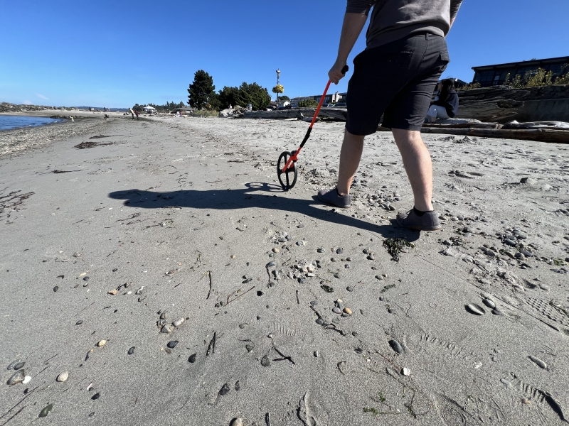 A person's back is seen as they walk down a sandy beach pushing a monitoring wheel on a sunny day.