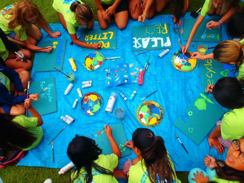 A group of students painting "Please Recycle" messages on a blue sign.