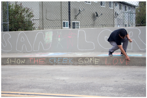 A student writes "show the creek some love" on the sidewalk.