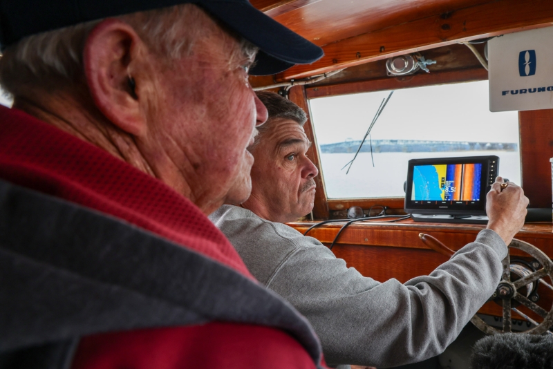 Two people sit inside the cabin of a boat facing towards a screen on a cloudy day.
